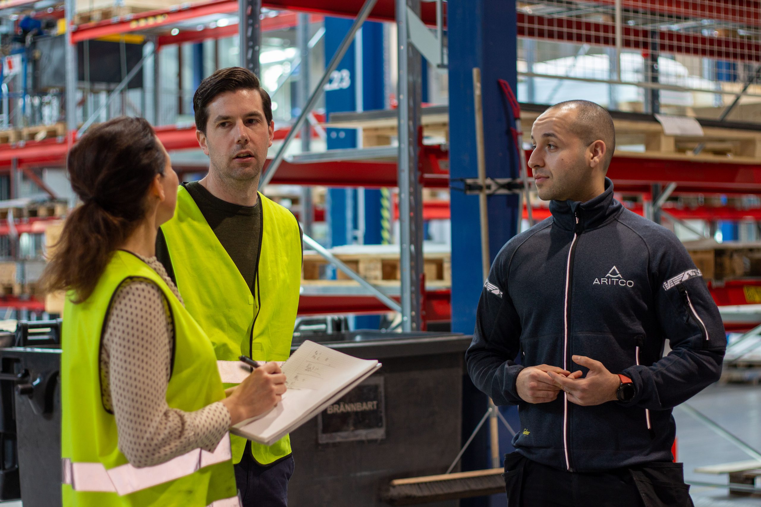 A woman doing an interview with paper and pencil with two men in  a production area